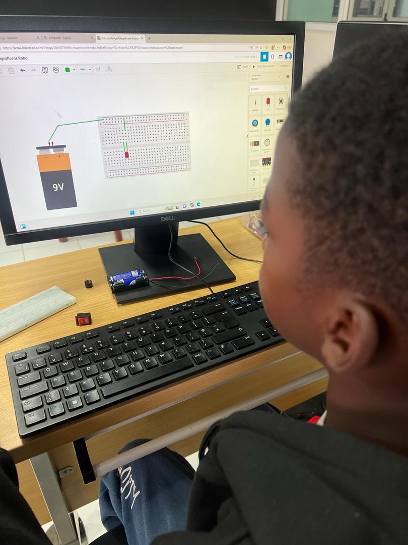 A child studies a Tinkercad circuit design on screen while building a 9V battery breadboard at CodeImpact Kampala